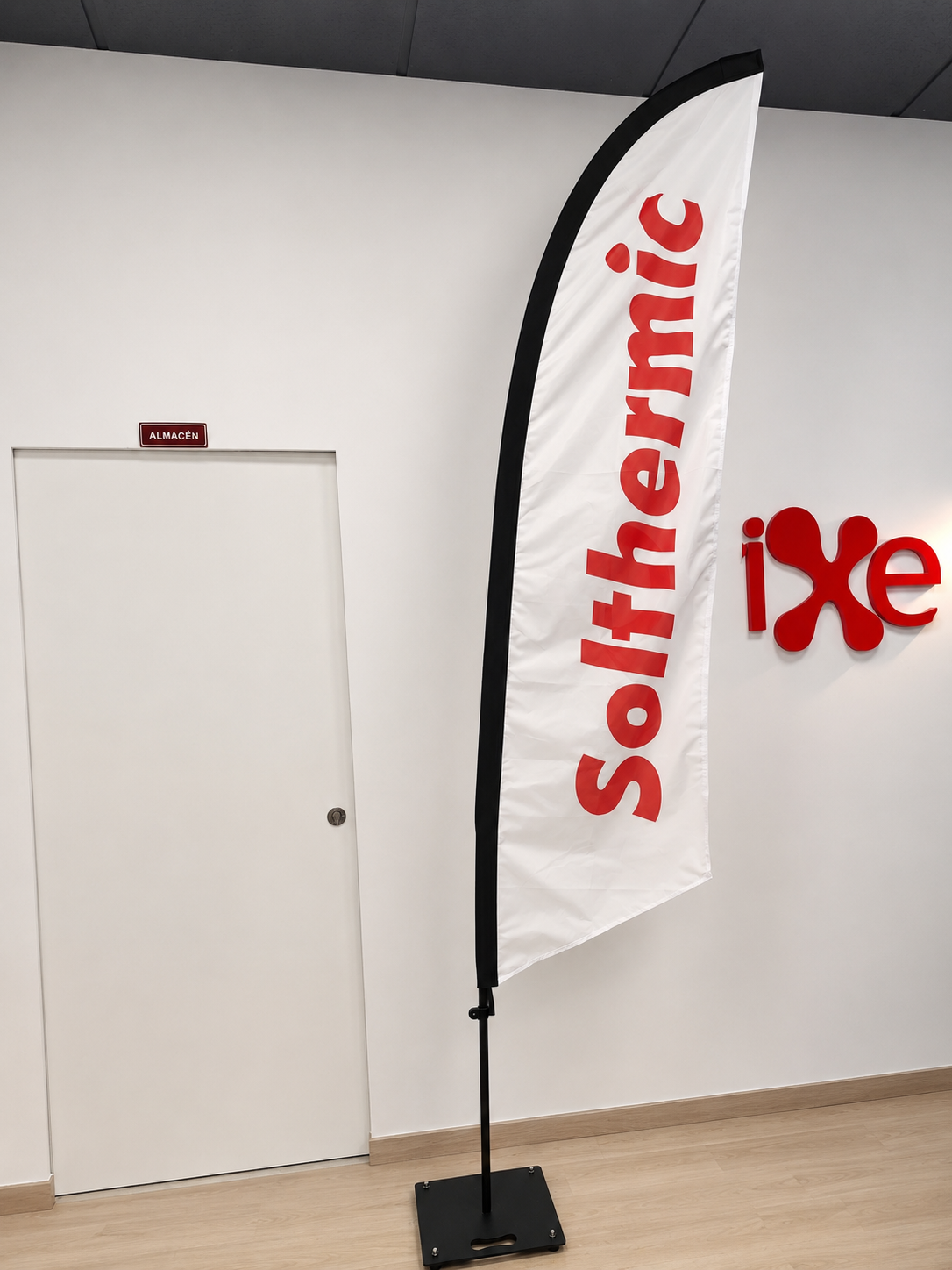 Bandera publicitaria Solthermic Bandera publicitaria blanca de Solthermic colocada en interior, con logotipo rojo sobre soporte vertical. Tipo surf 290cm
