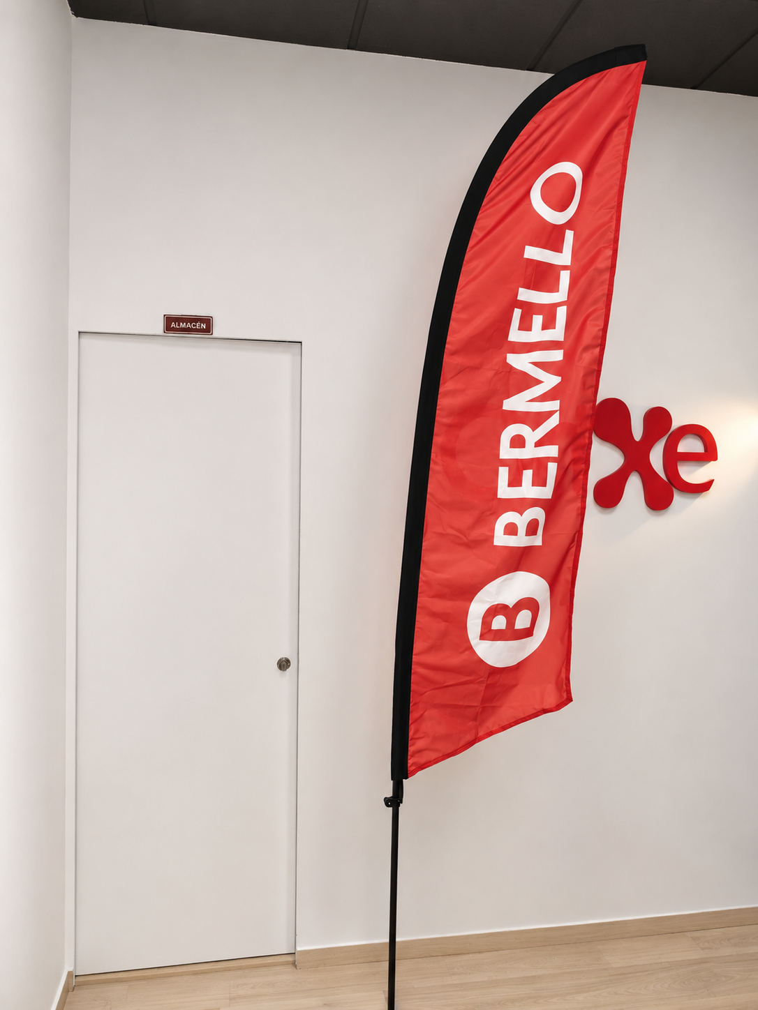Bandera publicitaria Bermello Bandera publicitaria roja de Bermello colocada en interior, con logotipo blanco sobre soporte vertical.